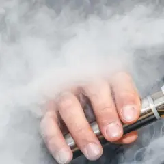 Mężczyzna zaciąga się e-papierosem, unosząc kłąb pary. Czy wapowanie jest szkodliwe? Dym otacza jego dłoń trzymającą urządzenie.