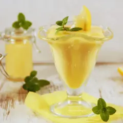 Pyszny, kremowy lemon curd w pucharku, ozdobiony plasterkiem cytryny i miętą. Obok dwa słoiczki z tym samym musem.