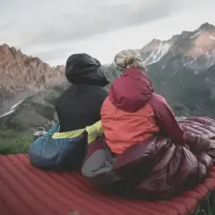 Dos personas sentadas en un colchón inflable, envueltas en sacos de dormir, disfrutan de un paisaje montañoso. El material vivac les permite acampar cómodamente.