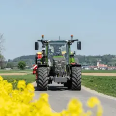Zielony traktor Fendt 516 jedzie drogą przez kwitnące pola rzepaku, w tle widać wieś.
