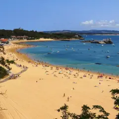 La **playa de los peligros** se llena de gente disfrutando del sol y el mar azul, con barcos anclados y edificios en la costa.
