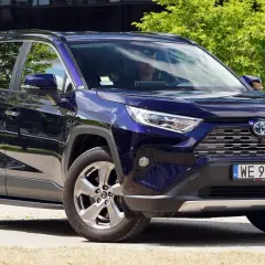 Elegancka, granatowa Toyota RAV4 5 generacji z przyciemnianymi szybami i chromowanymi felgami, zaparkowana na tle zieleni i nowoczesnego budynku.