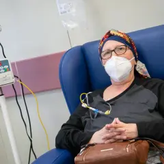 Mujer con mascarilla y pañuelo en la cabeza, sentada en una silla azul, recibiendo quimio en pastillas. Se le cae el pelo.