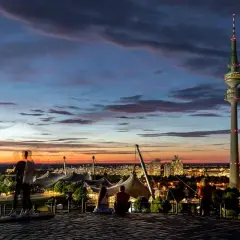 Tramonto a Monaco, Germania. La torre Olympiaturm domina il panorama, con le luci della città che iniziano ad accendersi, un'atmosfera da night club.