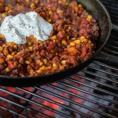 Chili con carne fumant dans une poêle sur un barbecue, garni de crème fraîche.