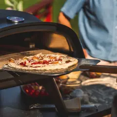 En pizza på muurikka är på väg in i en utomhusugn. Solen skiner och en person i blå skjorta står i bakgrunden.