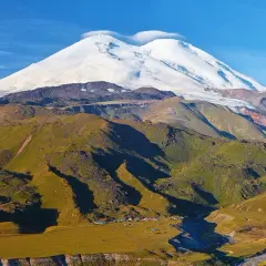 Elbrus szczyt w chmurach, otoczony zielonymi zboczami i obozem u podnóża.