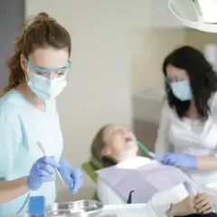 Dwie dentystki w maskach i okularach pracują przy pacjencie. Jedna z nich trzyma narzędzie, być może do usuwania pozostawionych szwów nierozpuszczalnych.
