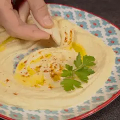 Un trozo de pan pita se sumerge en un cremoso hummus, adornado con aceite de oliva y perejil. Descubre qué es el tahini y cómo prepararlo.