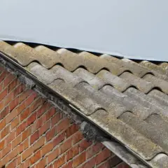 Stary dach pokryty eternitem, czyli płytami azbestowymi, z widoczną ceglaną ścianą pod nim.