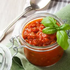 Tarro de cristal con salsa napolitana casera, decorado con hojas de albahaca fresca. Cuchara plateada y servilleta verde al lado.