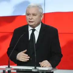 Jarosław Kaczyński w garniturze przemawia przy mównicy na tle flagi Polski.