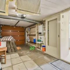 Un garage en cours de transformation en habitation. On y voit une porte de garage, un évier, une chaise en rotin et des éléments de rangement.