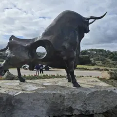 Escultura de toro en la plaza toros Valdemorillo. Paisaje rural con colinas verdes y cielo nublado.