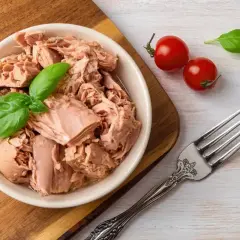Miseczka pełna tuńczyka w sosie własnym, ozdobiona listkami bazylii, obok dwa pomidorki i widelec.