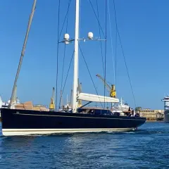 Il kauris iv yacht, scuro e imponente, naviga in un porto con gru e navi da crociera sullo sfondo.