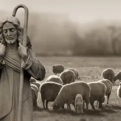 Kamienna rzeźba przedstawia, jak wygląda pasterz z owieczką na ramieniu, stojący na tle stada owiec pasących się na łące.