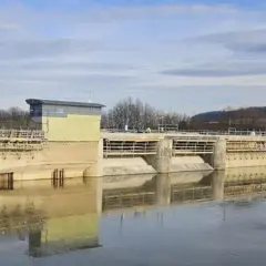 Betonowa zapora w Sieniawie w budowie, z widocznym budynkiem sterowni i rusztowaniami. Woda odbija konstrukcję.