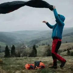 Osoba w niebieskiej kurtce i czerwonych spodniach rozkłada śpiwór turystyczny na tle górskiego krajobrazu.