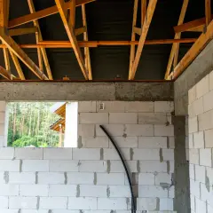 Budowa domu w toku. Widoczny wieniec stropowy, ściany z bloczków, drewniana więźba dachowa i widok na las.