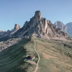 Un refugio alpino solitario en la ladera de una montaña, con un sendero que serpentea hacia él. El paisaje evoca la belleza de **Milano Cortina**, con picos imponentes y cielos despejados.