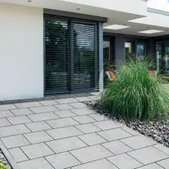 Nowoczesny podjazd z szarej kostki brukowej, otoczony kamieniami i zielenią, prowadzący do domu.