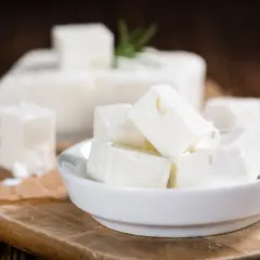 Cubos de queso de leche cruda en un cuenco blanco, con un bloque de queso y una ramita de romero al fondo.