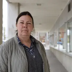 Jessica de Geer står utanför en byggnad med tegel och betong. Hon bär en grå jacka över en mönstrad skjorta och en stickad väst.