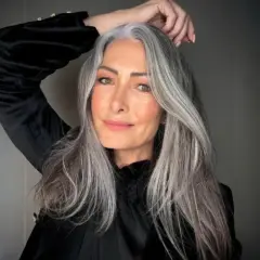 Mujer con cabello largo y ondulado, mostrando una hermosa integración de canas. Su mano acaricia su cabeza, resaltando la textura y el color natural.