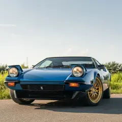 Niebieski De Tomaso Pantera z wysuwanymi światłami i złotymi felgami na tle zieleni i błękitnego nieba.