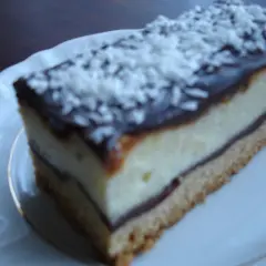 Szybkie ciasto z białym serem i budyniem, polane czekoladą i posypane wiórkami kokosowymi, na białym talerzyku.