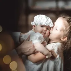 Une sœur embrasse tendrement son bébé, habillé pour baptiser son enfant. Lumières douces et ambiance chaleureuse.