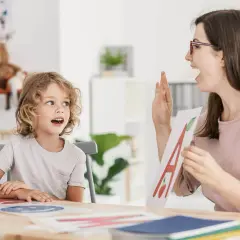 Chłopiec z kręconymi włosami uczy się liter, a nauczycielka pokazuje mu kartę z literą "A". To lekcja o samogłoskach.