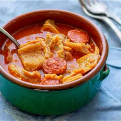 Un tazón de barro con callos a la madrileña, trozos de chorizo y un caldo rojizo. Una cuchara metálica está dentro.