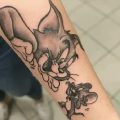 Czarno-biały tatuaż tom i jerry na przedramieniu. Kot Tom próbuje złapać myszkę Jerry, która ucieka.
