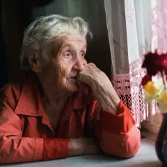 Eine ältere Frau blickt nachdenklich aus dem Fenster, ihre Hand am Kinn. Die Szene vermittelt ein Gefühl von **Angst vor Einsamkeit im Alter**.