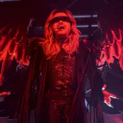 Bill Kaulitz z Tokio Hotel na koncercie w anielskich skrzydłach i błyszczącym stroju, w czerwonym świetle.