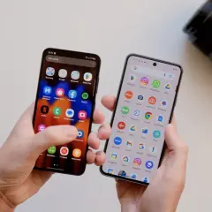 Dwa telefony w dłoniach, jeden z ikoną aparatu, drugi z ikoną aparatu. Który telefon ma lepszy aparat? Obok leży obiektyw aparatu.