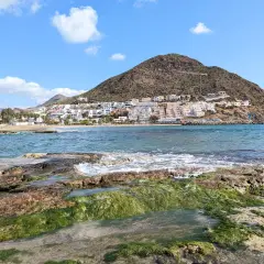 Costa de Cabo de Gata en octubre, con rocas cubiertas de algas verdes y el mar azul. Al fondo, el pueblo blanco se alza en la ladera de una montaña.