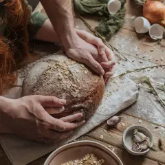 Dłonie obejmują bochenek chleba, jakby zastanawiając się, dlaczego chleb nie wyrasta. Na drewnianym stole mąka, jajka, cebula i czosnek.