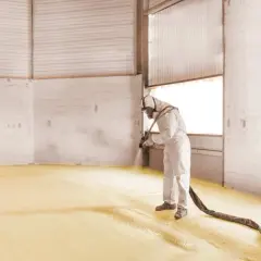 Un ouvrier en combinaison projette de la mousse polyuréthane sur le sol d'un entrepôt pour une isolation efficace.