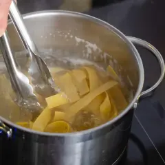 Szef kuchni dodaje szczypcami szeroki makaron pełnoziarnisty do gotującej się wody w garnku.