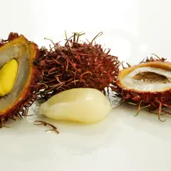 Dwa przekrojone i jeden cały, kolczasty, azjatycki owoc rambutan z białym miąższem i żółtym pestką.