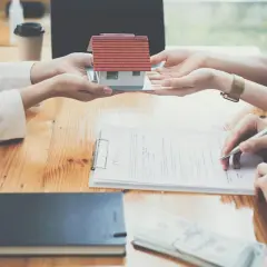 Dwie osoby dokonują zamiany nieruchomości o różnych wartościach bez dopłaty, przekazując sobie model domu i podpisując umowę.