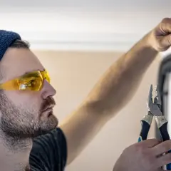 Elektryk w okularach ochronnych i czapce pracuje przy skrzynce elektrycznej, trzymając kombinerki. Zastanawia się, z której strony faza w gniazdku.