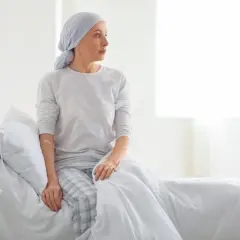 Kobieta w chuście na głowie siedzi na łóżku szpitalnym obok kroplówki.