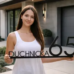 Uśmiechnięta kobieta trzyma tabliczkę z napisem "DUCHNICKA 65". To idealna cmg rejestracja dla nowego domu.