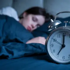 Frau schläft tief, während ein Wecker die Zeit anzeigt. Wie viel Schlaf braucht man mit 40? Die Antwort liegt im erholsamen Schlaf.
