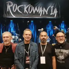 Czterech mężczyzn pozuje na tle sceny z banerem "ROCKOWANIA". Jeden z nich ma na sobie czerwoną kurtkę, inny dżinsową koszulę. W tle widać zespół i sprzęt. Idziemy do przodu dżem!
