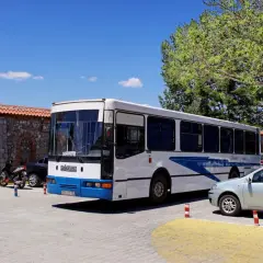 Biały autobus z niebieskimi pasami, oznaczony jako "Arbus", stoi zaparkowany obok kamiennego muru. To autobus z Macedonii, gotowy na kolejną podróż.